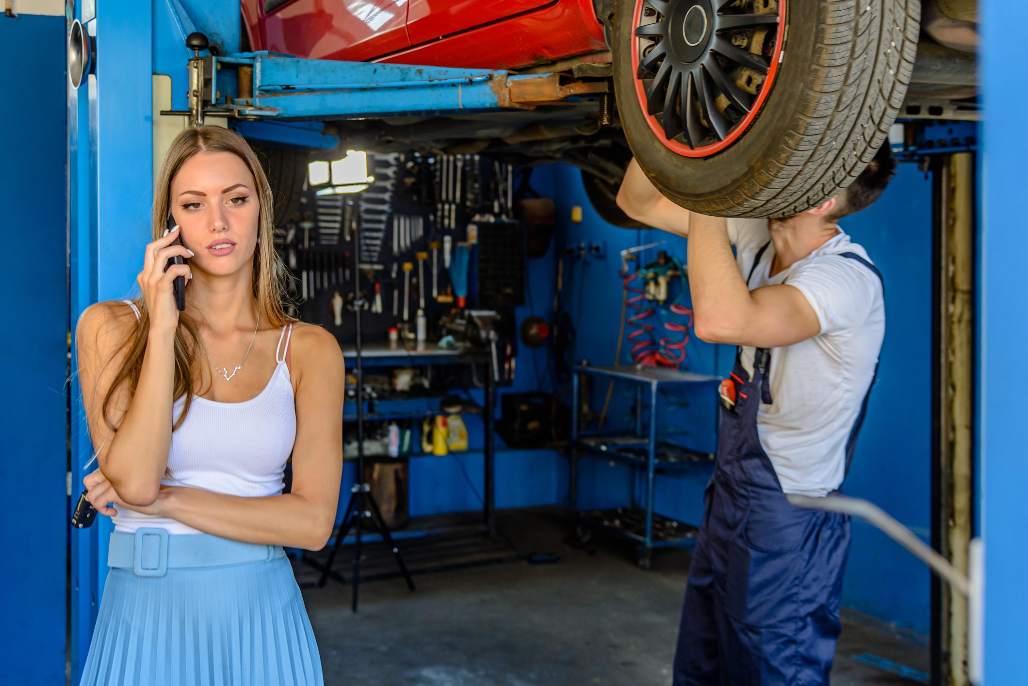Photo of an Experienced Car Mechanic who is Working Under the Car Using Tools for Work in Presence of a Young Female Customer Talking via the Mobile Phone.